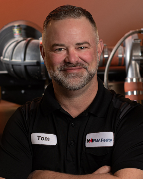Portrait of Tom Stieren, Maintenance Supervisor at NAI FMA Realty, standing in an industrial setting with arms crossed.